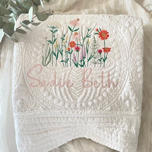 以下が含まれることがあります： 白いキルティングのベビーブランケットに、花柄の刺繍とピンク色の刺繍で「Sadie Beth」という名前が入っています。