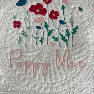 May include: White quilted baby blanket with embroidered pink flowers and green leaves. The blanket has the name "Poppy Mae" embroidered in pink cursive lettering.