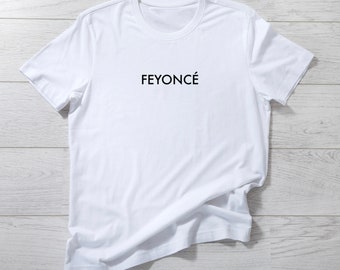 Può includere: Maglietta bianca con la scritta "FEYONCÉ" stampata in nero sul davanti.