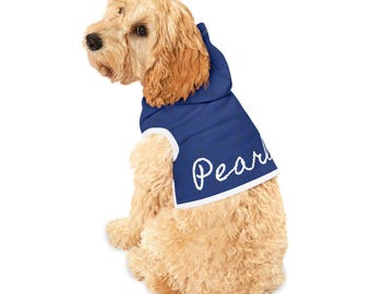 May include: A blue dog hoodie with a white trim and the name "Pearl" embroidered on the front. The hoodie has a hood and is worn by a light brown dog.