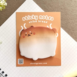 May include: A square sticky note pad featuring a Shiba Inu dog design, styled as a loaf of bread. The card has the text "sticky notes shiba bread" in white. The background is a light orange colour.