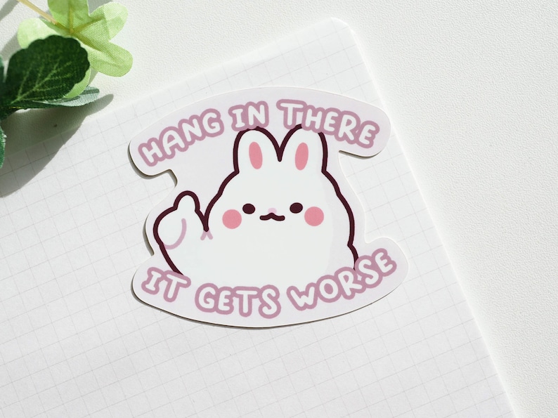 May include: A white bunny sticker with pink accents, giving a thumbs-up. The sticker has the text "Hang in there" above the bunny and "It gets worse" below. The sticker is on a white grid paper.