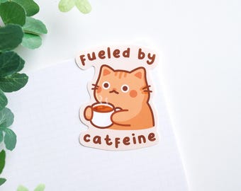Könnte beinhalten: Ein Aufkleber mit einer orangefarbenen Comic-Katze, die eine Tasse Kaffee hält. Der Text "fueled by caffeine" steht unter der Katze.