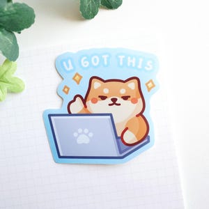 Peut inclure: Autocollant représentant un chien Shiba Inu avec un ordinateur portable. Le chien est orange et blanc, faisant un signe de pouce levé. L'autocollant porte le texte "U GOT THIS" en bleu clair, avec des accents de diamant. L'ordinateur portable est bleu clair avec une empreinte de patte.