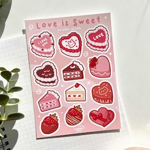 May include: A sticker sheet with a pink background featuring various Valentine's Day themed designs. The sheet includes hearts, cakes, and strawberries, all with a pink and red colour scheme. The text "Love is Sweet" is printed at the top of the sheet.