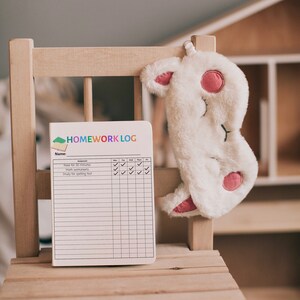 May include: A white wooden chair with a white and pink sleep mask hanging on the back. A white notebook with the text "HOMEWORK LOG" is on the seat. The notebook has a checklist with the assignments "Read for 30 minutes", "Math worksheets", and "Study for spelling test".