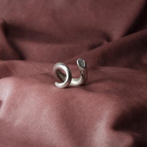 Peut inclure: Un ear cuff argent&eacute; avec un motif en spirale. Le bijou s'enroule autour de l'oreille, avec une spirale arrondie &agrave; une extr&eacute;mit&eacute; et une extr&eacute;mit&eacute; droite et ouverte. Le bijou est pos&eacute; sur un fond bordeaux.