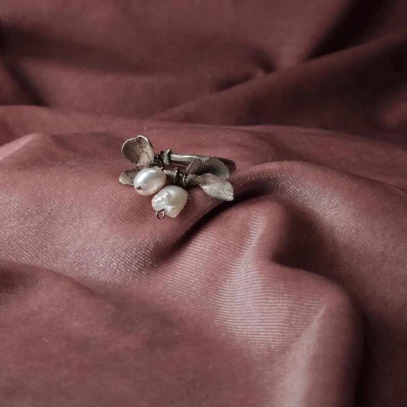 Peut inclure: Une bague argent&eacute;e orn&eacute;e d'un motif floral et de trois perles blanches en forme de poire. La bague pr&eacute;sente une esth&eacute;tique d&eacute;licate inspir&eacute;e de la nature, avec des d&eacute;tails de feuilles et de p&eacute;tales. La bague est pos&eacute;e sur un tissu rose tendre.