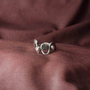 Peut inclure: Une bague argent&eacute;e au design unique. La bague pr&eacute;sente un &eacute;l&eacute;ment central circulaire avec un petit motif plat, flanqu&eacute; de formes courbes et abstraites. La bague est pos&eacute;e sur un fond bordeaux.