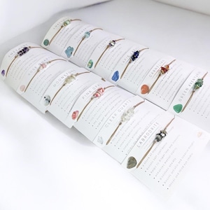 May include: Assortment of ten gemstone bracelets on cards with descriptions of each stone. The bracelets are made with brown string and feature various gemstones, including amethyst, rose quartz, moonstone, fluorite, lapis lazuli, carnelian, green aventurine, pink opal, clear quartz, and labradorite.