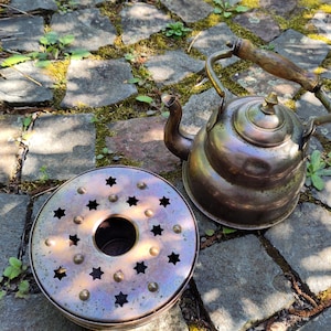 Könnte beinhalten: Ein antiker Kupfer-Teekessel mit einem Holzgriff und einem passenden Kupferständer mit sternförmigen Ausschnitten. Der Kessel und der Ständer stehen auf einer Steinplatte.