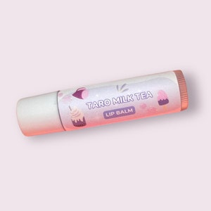 May include: A pink and white lip balm tube with a glittery finish. The label features a cartoon illustration of a taro milk tea drink and the text "Taro Milk Tea Lip Balm".