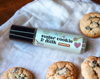 May include: A small glass bottle of sugar cookie and milk scented perfume oil. The bottle has a colorful label with the text "sugar cookie & milk perfume oil". The bottle is sitting on a white cloth with four sugar cookies.