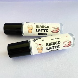May include: Two clear glass roller bottles with black caps. Both bottles have white labels with a cartoon image of a coffee drink and the text "Inspired Collection Bianco Latte Perfume Oil".