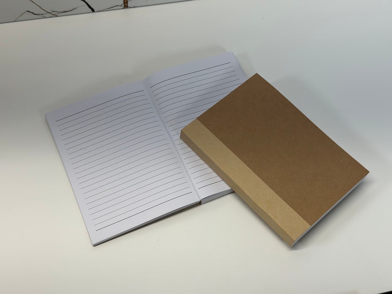 Puede incluir: Dos cuadernos, uno con una cubierta blanca y p&aacute;ginas rayadas, y el otro con una cubierta marr&oacute;n y un lomo marr&oacute;n.