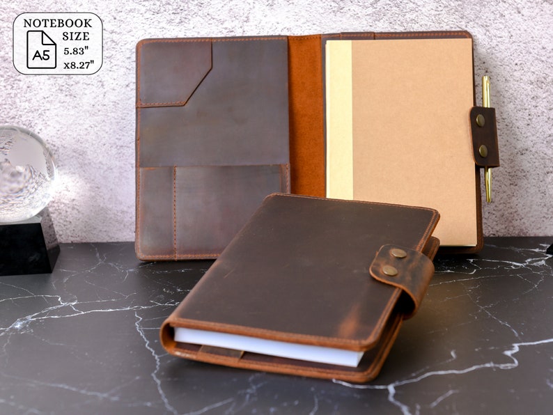 May include: A brown leather notebook cover with a pen holder and a flap that closes with a button. The cover is open to reveal a blank notebook page. The notebook size is A5, 5.83 inches by 8.27 inches.