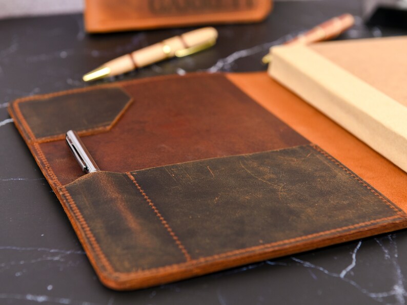 May include: A brown leather notebook cover with a pen holder and a dark brown leather flap. The cover has a distressed look and is made of high-quality leather.