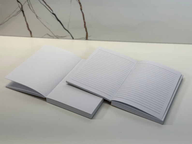 Puede incluir: Dos cuadernos en blanco, uno abierto para revelar p&aacute;ginas rayadas, el otro cerrado. Ambos cuadernos son blancos sobre un fondo blanco.