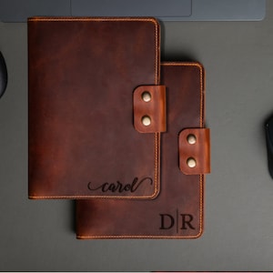 May include: Two brown leather journals with personalized engravings. The top journal has the name "carol" engraved in cursive script. The bottom journal has the initials "D|R" engraved in a block font.