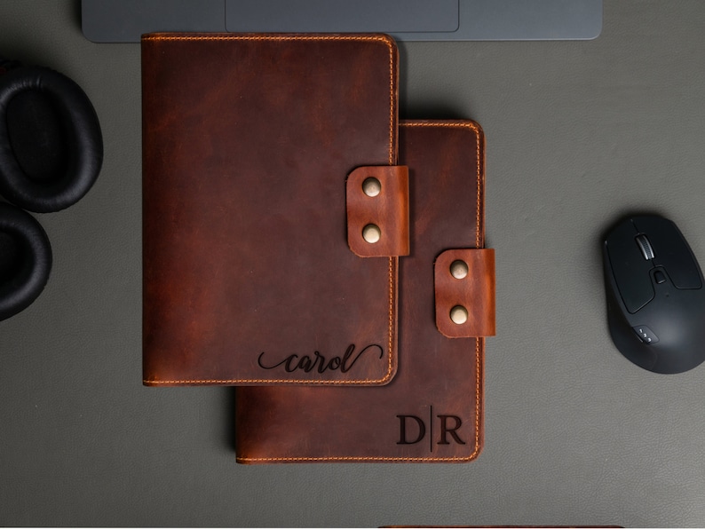 May include: Two brown leather journals with personalized engravings. The top journal has the name "carol" engraved in cursive script. The bottom journal has the initials "D|R" engraved in a block font.