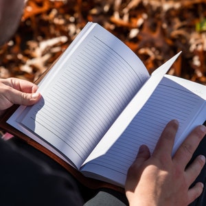 May include: A person sitting outdoors, reading a blank, lined journal with a brown leather cover.