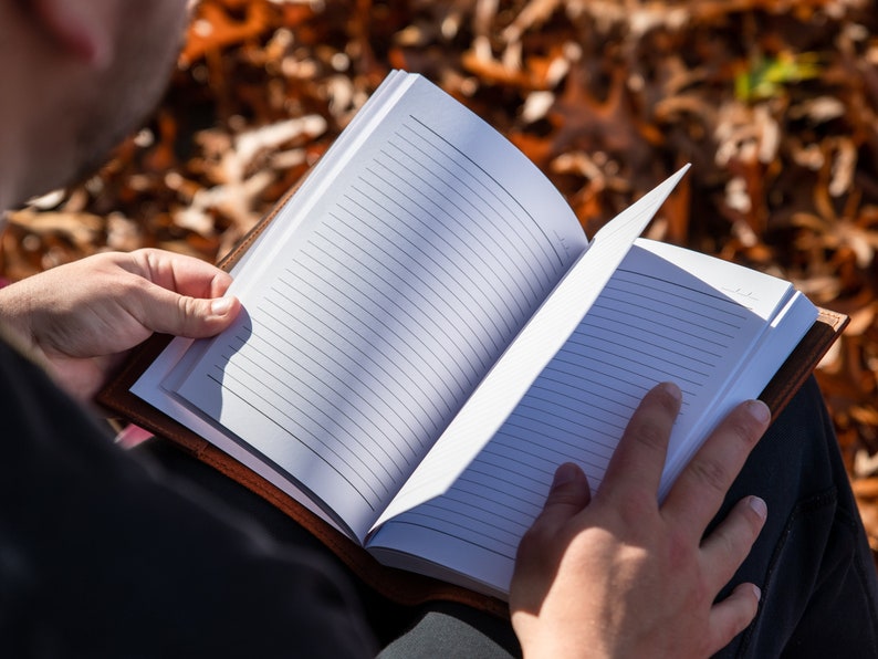 May include: A person sitting outdoors, reading a blank, lined journal with a brown leather cover.