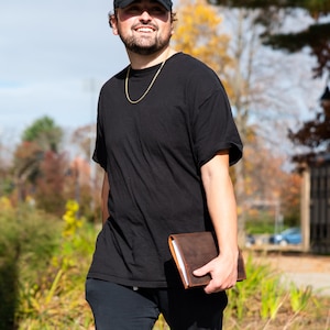 May include: A person wearing a black t-shirt, a black baseball cap, and black pants is holding a brown leather journal.
