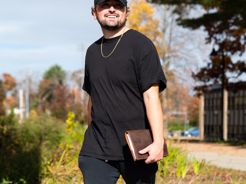 May include: A person wearing a black t-shirt, a black baseball cap, and black pants is holding a brown leather journal.