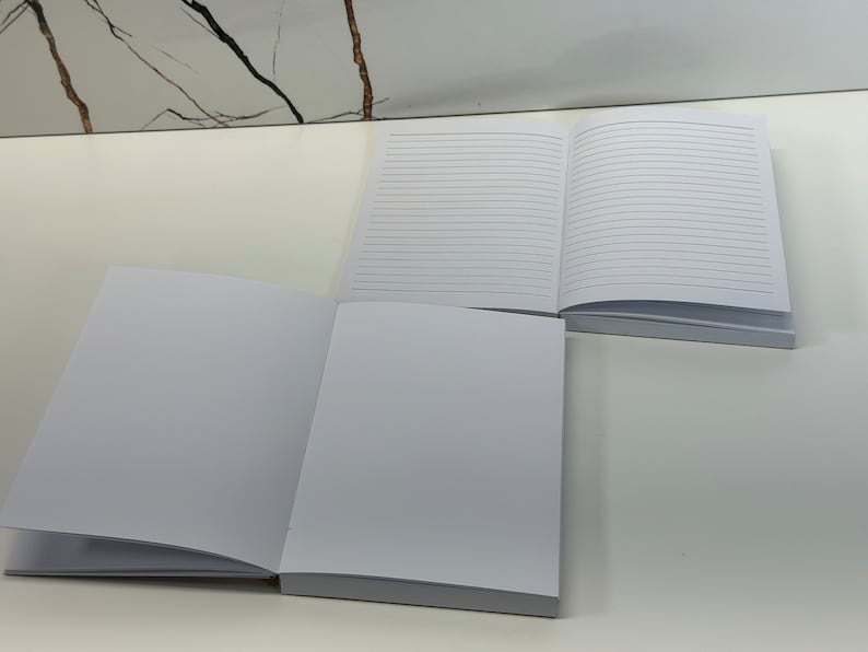 Puede incluir: Dos cuadernos en blanco con cubiertas blancas y p&aacute;ginas rayadas. Los cuadernos est&aacute;n abiertos y colocados planos sobre una superficie blanca.