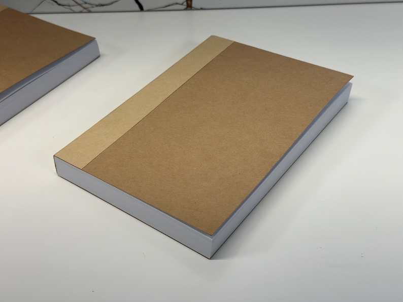 Puede incluir: Un cuaderno marr&oacute;n con una encuadernaci&oacute;n blanca y una cubierta de papel marr&oacute;n. El cuaderno est&aacute; cerrado y sobre una superficie blanca.