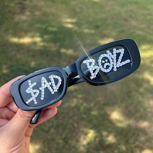 May include: Black sunglasses with rectangular frames. The left lens features the word "$AD" in rhinestone lettering, and the right lens displays "BOIZ" with a sad face emoji, also in rhinestones. The sunglasses are held against a blurred green background.