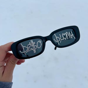 Könnte beinhalten: Schwarze rechteckige Sonnenbrille mit Strasssteinen, die "babe" und "blond" buchstabieren. Die Sonnenbrille wird vor einem verschneiten Hintergrund gehalten.