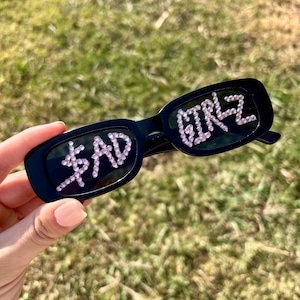 May include: Black rectangular sunglasses with the words "BAD GIRLZ" spelled out in pink rhinestones. The sunglasses are held up against a background of green grass.