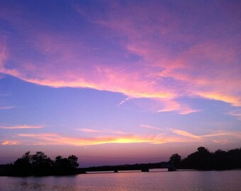 Könnte beinhalten: Eine malerische Aussicht auf einen See bei Sonnenuntergang mit einem rosa und violetten Himmel. Das Wasser ist ruhig und spiegelt die Farben des Himmels wider. Silhouettierte Bäume säumen das Ufer.