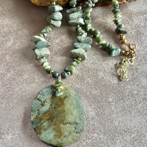 May include: A handcrafted necklace featuring a large, oval pendant with green and brown tones. The necklace is composed of small, green stone beads and larger, stacked stone elements. A gold-colored clasp secures the necklace.