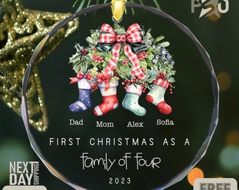 May include: A clear, round Christmas ornament with a festive design. It features a wreath with holly and berries, four stockings, and the text "First Christmas as a Family of Four 2023." The ornament is hanging from a gold ribbon.