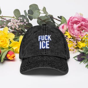 May include: A black, distressed baseball cap with the words "FUCK ICE" in white and blue block letters. The hat is surrounded by colourful flowers, including pink peonies, yellow daffodils, and red tulips. The cap has a curved brim and a classic six-panel design.