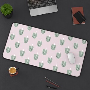 May include: A rectangular pink desk mat with a repeating pattern of green bubble tea cups. The mat has a black border and is shown with a laptop, mouse, and other desk accessories.