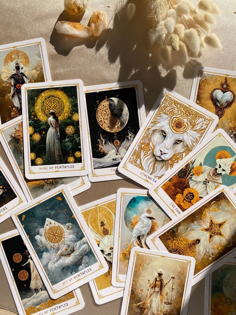 May include: A collection of tarot cards with gold and white illustrations, featuring images of a lion, a crescent moon, and figures in ornate attire. The cards are scattered on a beige surface, with the words "Strength", "The Fool", and "Ace of Pentacles" visible.