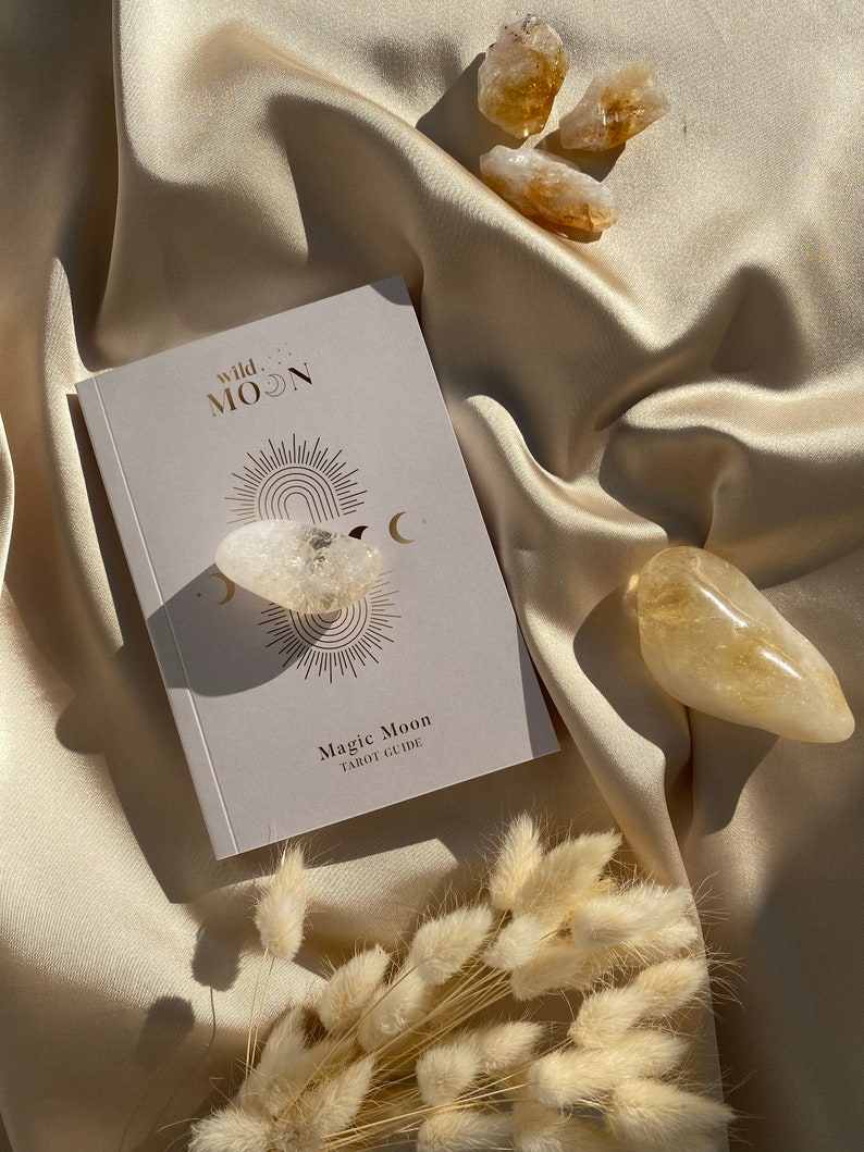 May include: A tarot guide titled "Magic Moon" with gold foil accents, a clear quartz crystal, and several citrine crystals. The items are arranged on a cream-colored satin fabric, with dried bunny tail grass in the foreground.