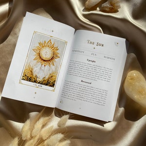 May include: An open book with a gold-colored sun illustration and the text "The Sun" on the pages. The book is on a satin surface with dried flowers and crystals.