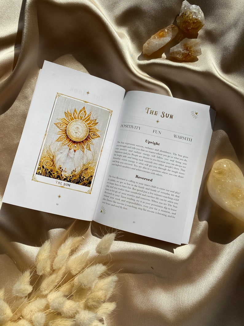 May include: An open book with a gold-colored sun illustration and the text "The Sun" on the pages. The book is on a satin surface with dried flowers and crystals.
