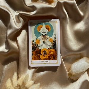 May include: A tarot card with a detailed illustration of a figure in white robes and a mask, surrounded by yellow flowers and a blue halo. The card reads "THE FOOL" in a gold font. The card is on a satin surface.