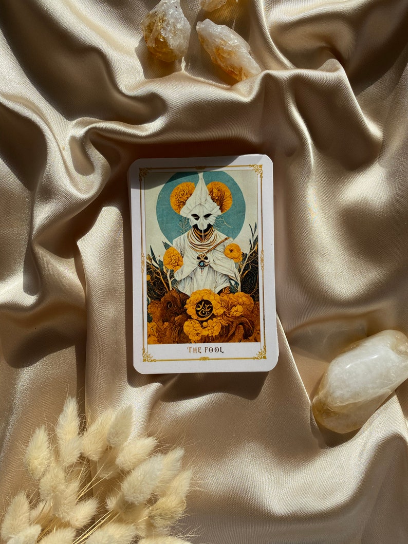 May include: A tarot card with a detailed illustration of a figure in white robes and a mask, surrounded by yellow flowers and a blue halo. The card reads "THE FOOL" in a gold font. The card is on a satin surface.