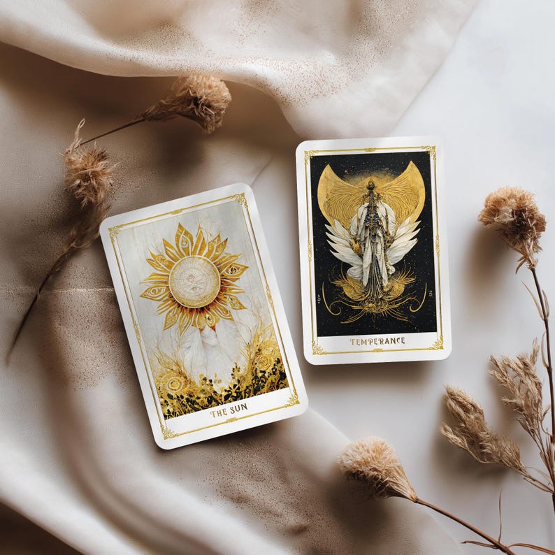 May include: Two tarot cards with gold and white designs. One card features a sun with eyes and the text "THE SUN". The other card shows a figure with wings and the text "TEMPERANCE". The cards have gold borders and are set on a white surface.