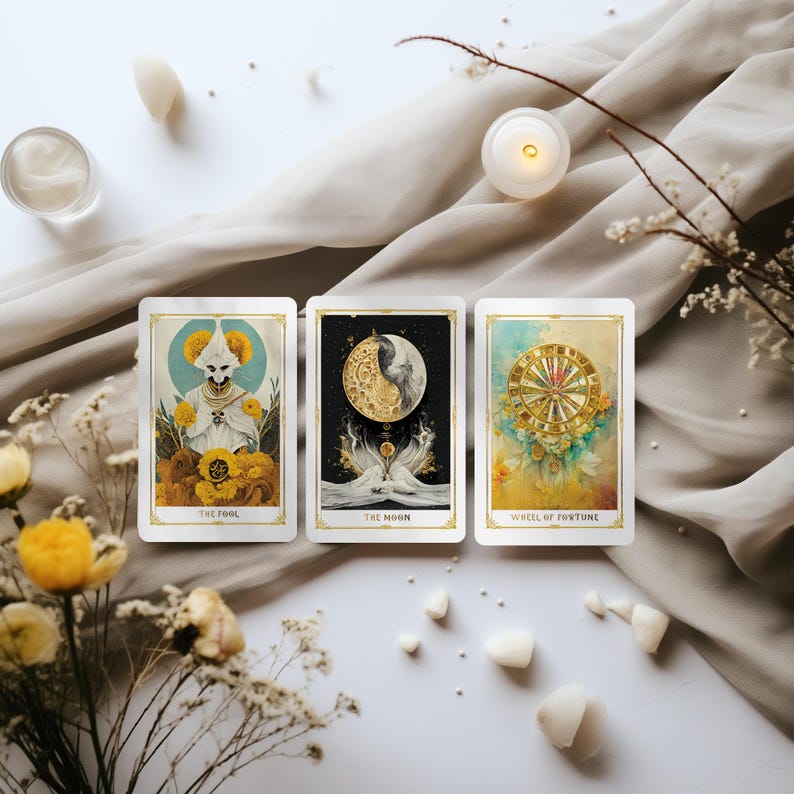 May include: Three tarot cards with gold borders are displayed, featuring illustrations of "The Fool," "The Moon," and "Wheel of Fortune." The cards are arranged on a white surface with decorative elements like flowers, candles, and crystals.