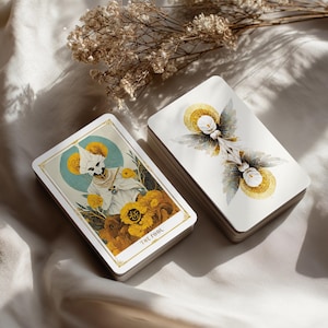 May include: Two decks of tarot cards. One card shows a figure in white robes and a skull face, with the text "THE FOOL". The other deck shows a symmetrical design with white flowers and gold accents. The cards are on a white cloth.