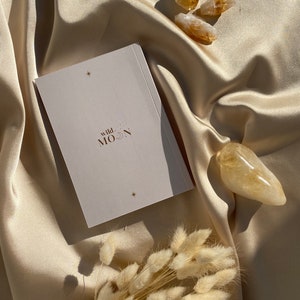 May include: A still-life composition featuring a deck of cards with the text "wild MOON" and star accents, alongside several golden citrine crystals and dried bunny tail grass. The items rest on a cream-colored satin fabric.