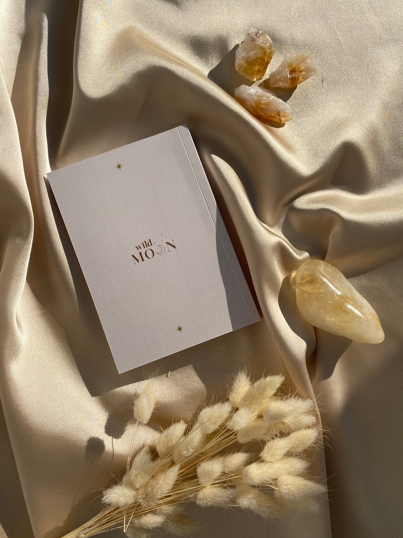 May include: A still-life composition featuring a deck of cards with the text "wild MOON" and star accents, alongside several golden citrine crystals and dried bunny tail grass. The items rest on a cream-colored satin fabric.