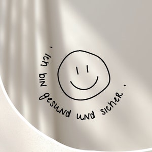 Könnte beinhalten: Ein weißer runder Spiegel mit einem schwarzen Smiley und dem Text "Ich bin gesund und sicher." drumherum.